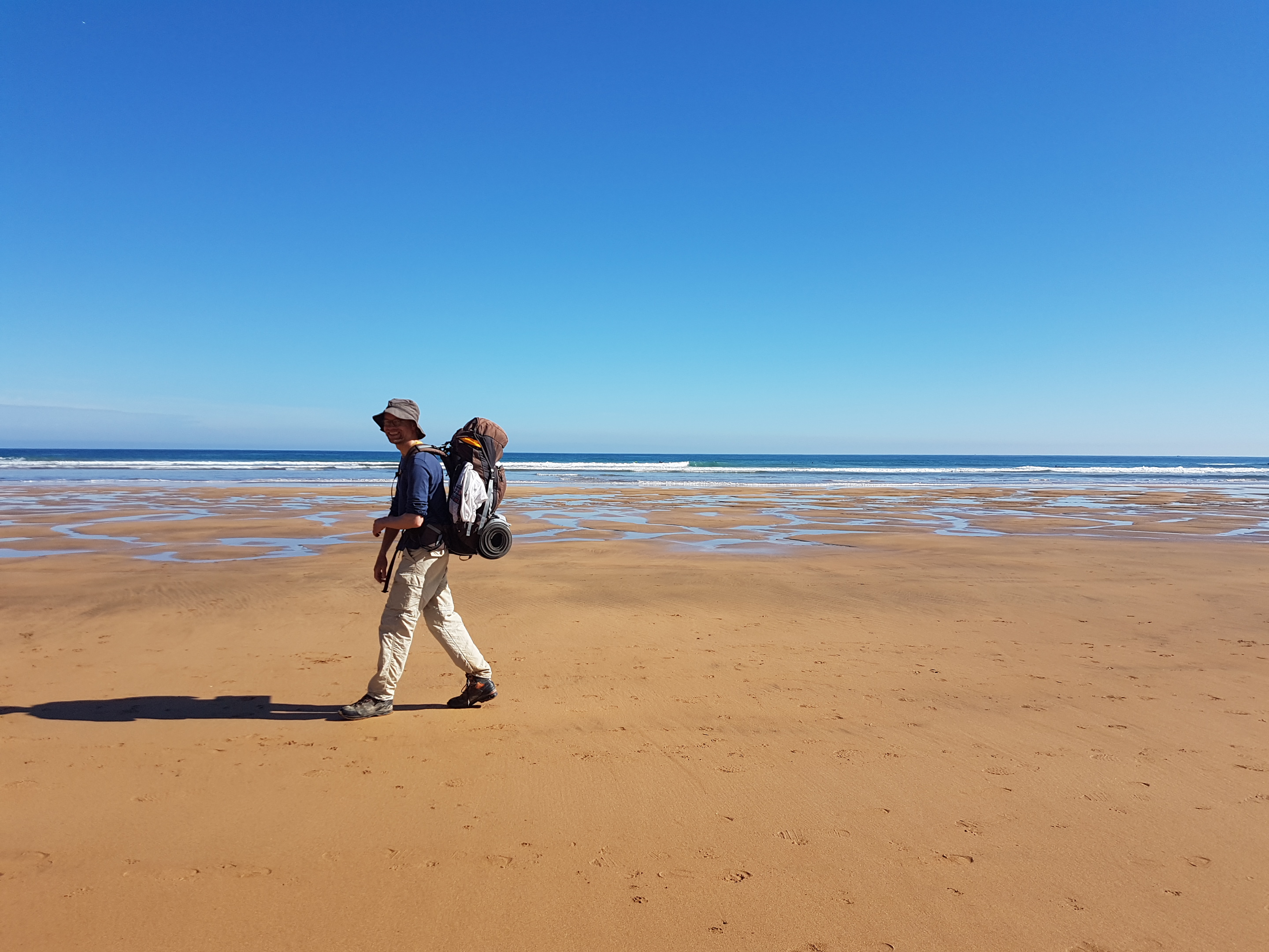 Person beim Wandern mit großem Rucksack auf dem Rücken und Mütze auf dem Kopf an einem Sandstrand